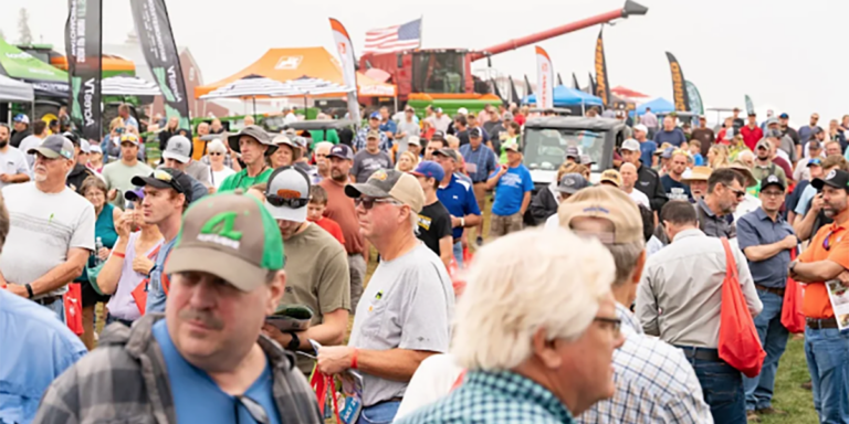 Crowd of people at an outdoor farm equipment trade show with tents and machinery