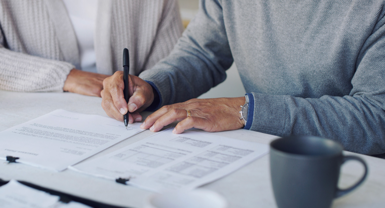 Person signing a document on a clipboard next to another person and a mug