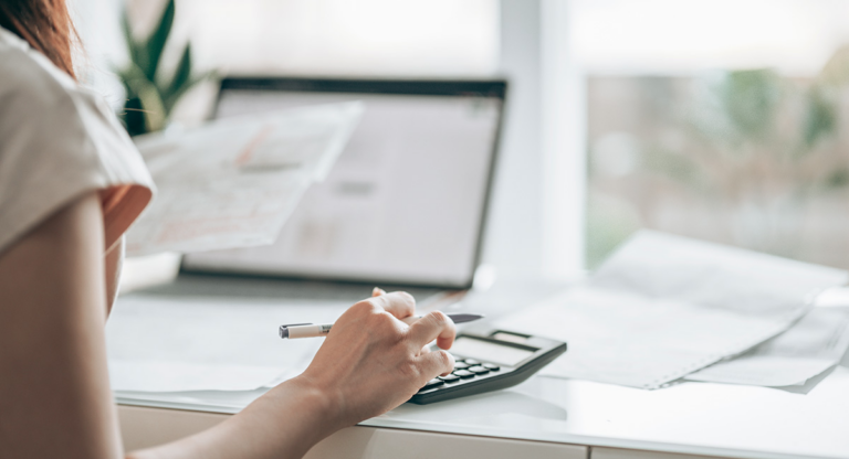 Woman using a calculator while holding a document at a desk with a laptop