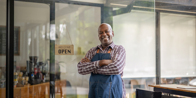 Man wearing an apron standing in a shop doorway next to a sign reading Welcome Open