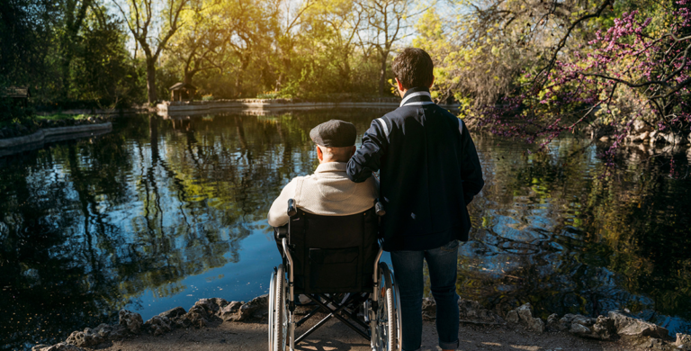 Man standing beside an older man in a wheelchair, both looking out over a pond