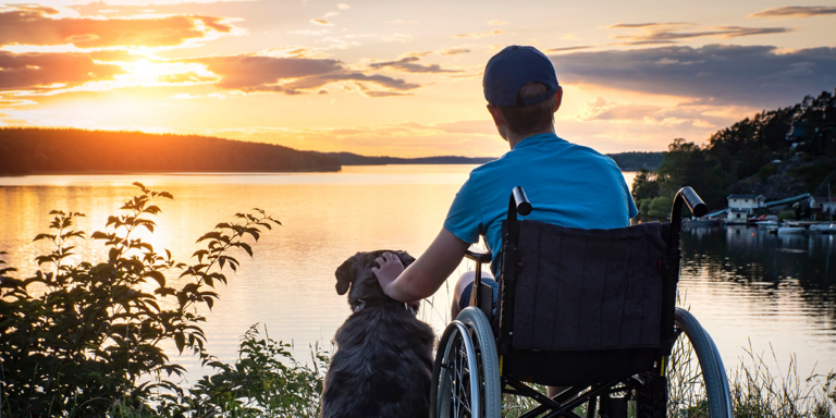 Boy in a wheelchair petting a dog while looking out over a lake at sunset