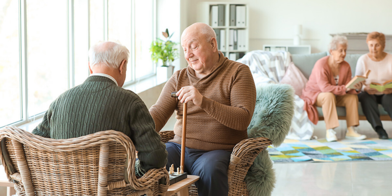 Elderly men playing chess in a common room while two women read on a couch behind them