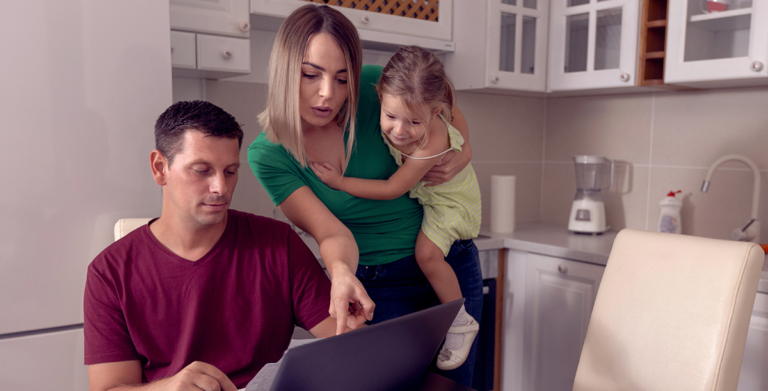 Woman holding a child and pointing at a laptop while a man looks at the screen in a kitchen