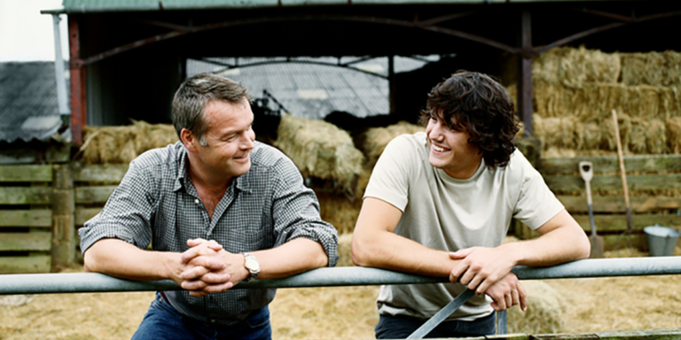Man and young man leaning on a fence rail talking on a farm