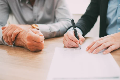 Younger person writing on a document at a table next to an older person