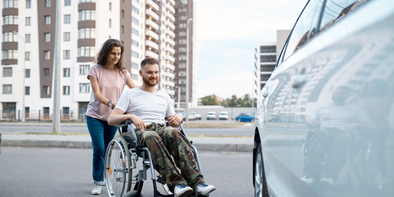 Woman pushing a man in a wheelchair toward a car on a city street