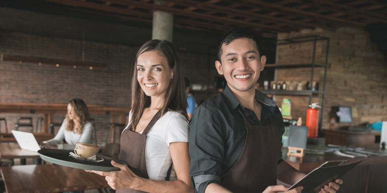 Woman and man wearing aprons in a cafe, one holding a tray with a coffee cup and the other holding a menu