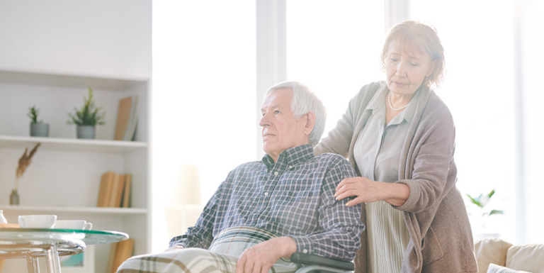 Older woman standing behind an older man seated in a wheelchair with her hand on his shoulder