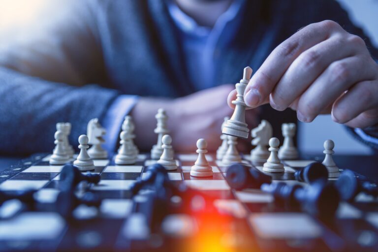 Person moving a white chess piece across a board with fallen pieces around it